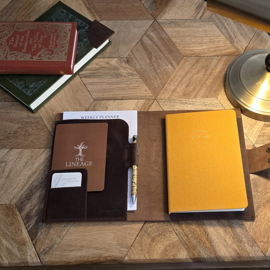 Top-down lifestyle view of brown leather Notebook Cover arranged with books and a desk lamp