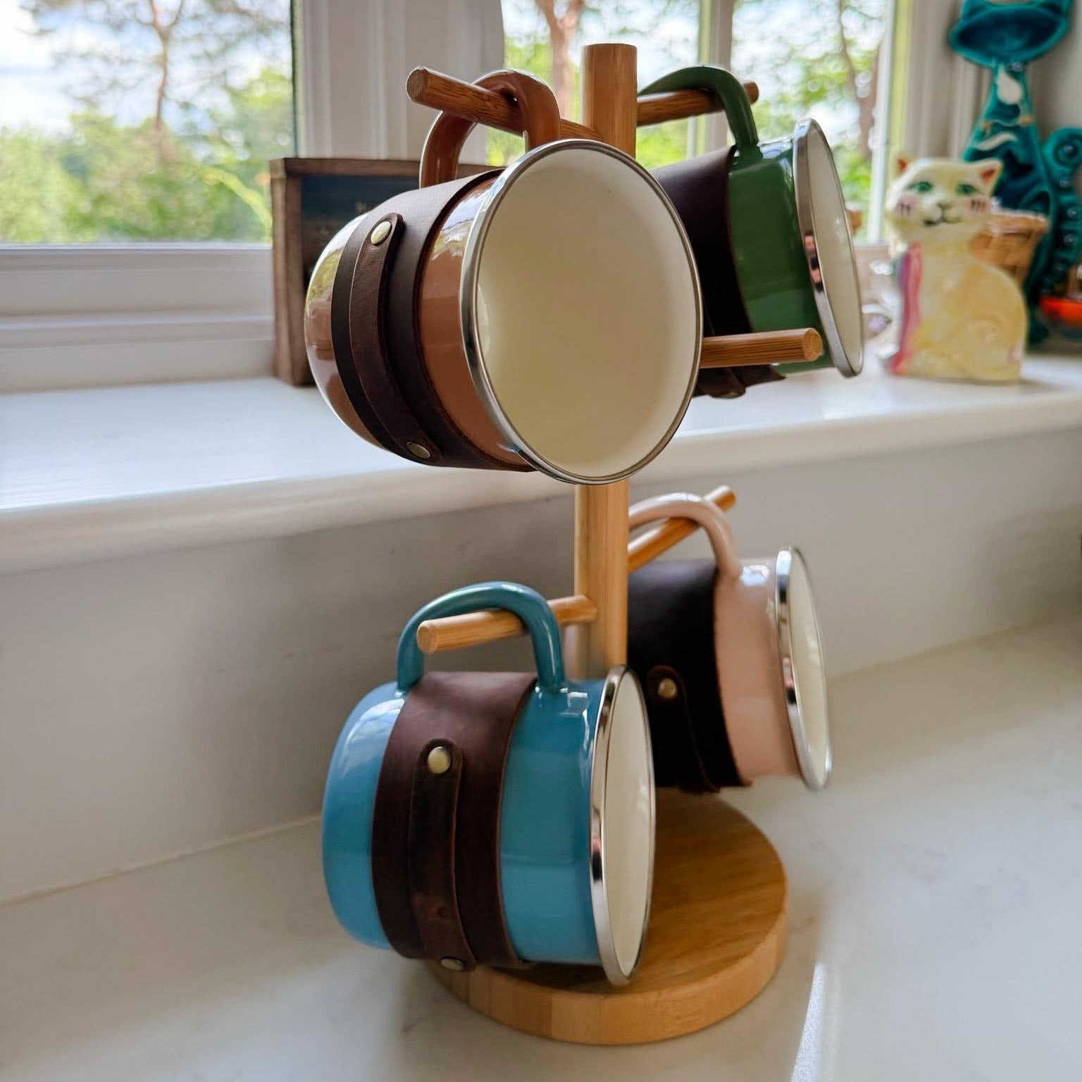 Enamel mugs in different colours with brown top grain crazy leather sleeves displayed on a wooden mug stand