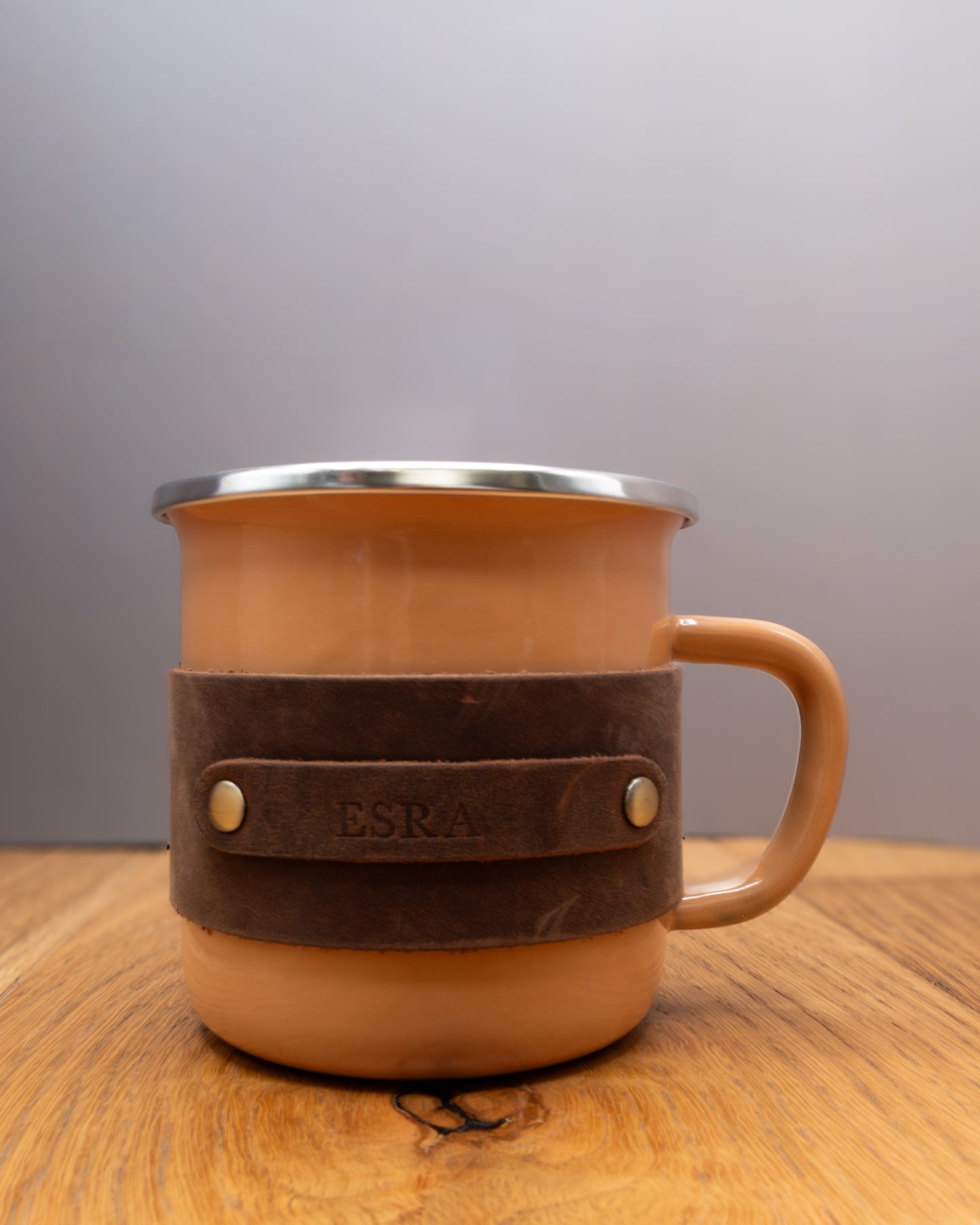 Personalised brown enamel mug with a brown top grain crazy leather sleeve engraved with a name on a wooden surface