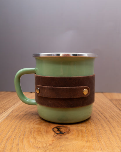 Green enamel mug with a brown top grain crazy leather sleeve on a wooden surface